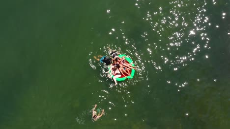 Aerial,-Group-of-friends-playing,-shoving-each-other-off-raft-into-lake