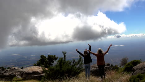 Two-women-with-hands-in-air-enjoy-view-at-Mariepskop,-from-behind