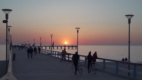 People-Walking-on-Palanga-Pier-on-Late-Evening-with-Sun-Setting-in-Sky-and-Flags-Waving-in-Wind