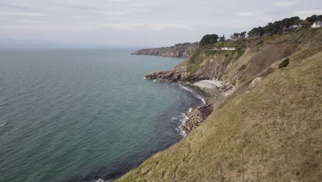 Aerial-Reveal-Of-A-Tiny-Hidden-Beach-In-Howth,-Dublin,-Ireland-With-Faraway-View-Of-Bailey-Cottage-On-The-Cliff