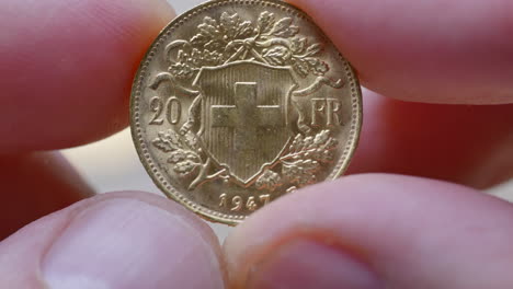 Macro-shot-of-male-finger-holding-swiss-golden-twenty-franc-coin-with-Helvetia-and-Swiss-Cross
