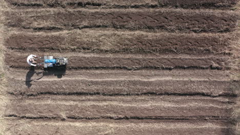 AERIAL---Man-working-a-field-with-a-rototiller,-agriculture,-rising-wide-shot