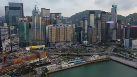 Aerial-View-of-Hong-Kong-Central-Waterfront-Corporate-Buildings,-Evening-Traffic-and-Construction-Site-on-Promenade,-Revealing-Drone-Shot