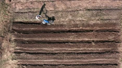 AERIAL---Man-working-a-field-with-a-rototiller,-agriculture,-wide-shot-tilt-up