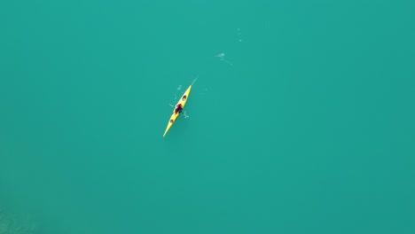 Woman-kayaking-through-clean-vibrant-green-glacier-water-in-yellow-kayak---Static-birdseye-aerial-view-with-kayak-passing-through-frame---Lovatnet-Lake-in-Loen-Nordfjord-Norway