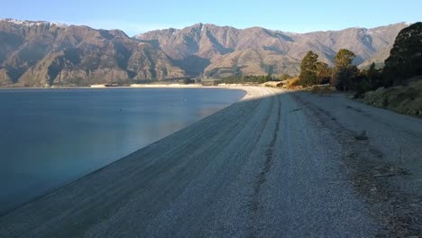 Wide-smooth-aerial-along-the-beach-at-the-south-of-Lake-Hawea-on-New-Zelands-South-Island,-a-popular-swimming-spot-with-locals-and-tourists-alike