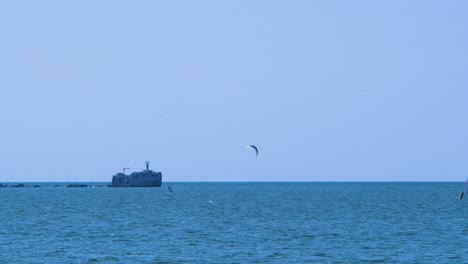 Lone-kiteboarder-sailing-in-distance,-concrete-port-pier-in-background,-sunny-summer-day,-calm-Baltic-sea-Karosta-beach,-medium-distant-shot
