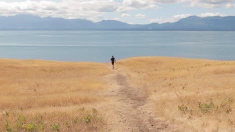 Man-running-down-the-golden-Helliwell-meadow-towards-the-ocean-with-the-Rocky-Mountains-in-the-background-on-Hornby-Island-in-British-Columbia,-Canada