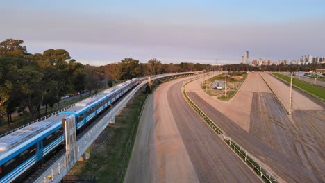 Incoming-Mitre-train-running-on-railroad-with-downtown-cityscape-along-the-skyline-at-sunset