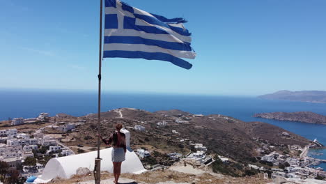 Vista-Aérea-Desde-La-Cima-De-La-Isla-De-Ios-En-Grecia,-Mujer-Y-Bandera-Griega