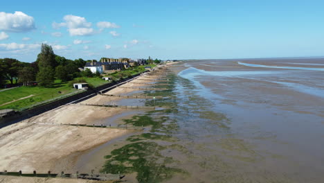 A-drone-flight-over-a-beach-with-old-wooden-groynes-and-mudflats,-with-homes-behind-a-sea-wall-on-the-left,-on-tidal-estuary-at-low-tide-on-a-clear-and-hot-summer-day