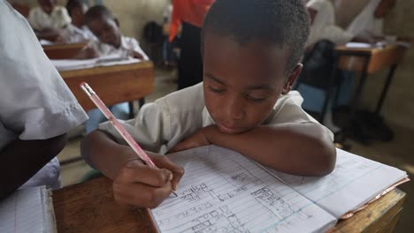 Trauriger-Afrikanischer-Schwarzer-Junge,-Der-In-Der-Schule-Mit-Kindern-In-Der-Hintergrundklasse-Schreibt-Und-Lernt