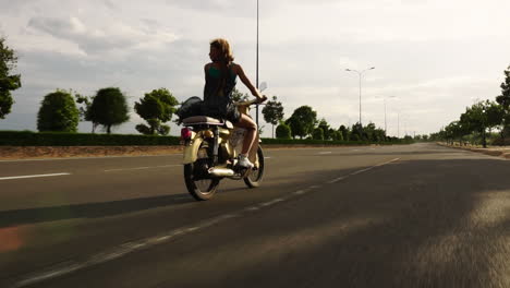 The-beautiful-young-blonde-caucasian-girl-is-riding-on-an-empty-highway-on-a-yellow-scooter---dolly-shot-from-behind