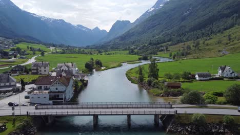 Tourists-traveling-through-Olden-city-center-over-concrete-bridge---closeup-forward-moving-aerial---Oldedalen-mountain-valley-raging-in-background---Norway