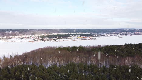 Frozen-lake-and-town-in-horizon-in-winter-season-during-snowfall,-aerial-drone-view
