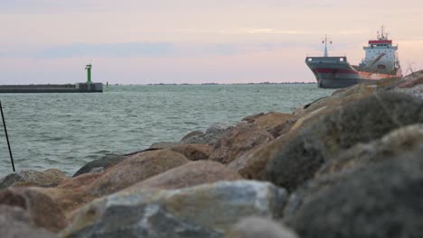 Large-gray-cargo-ship-entering-the-Port-of-Liepaja-in-calm-sunny-summer-evening,-stone-pier-in-foreground,-waves-splashing,-distant-medium-low-angle-shot