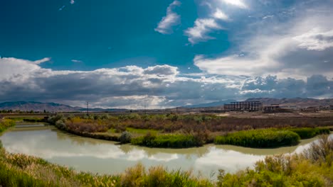Cloudscape-over-a-river-with-the-sky-reflection-on-the-water-and-a-highway-and-construction-project-by-the-distant-mountains---zoom-out-time-lapse
