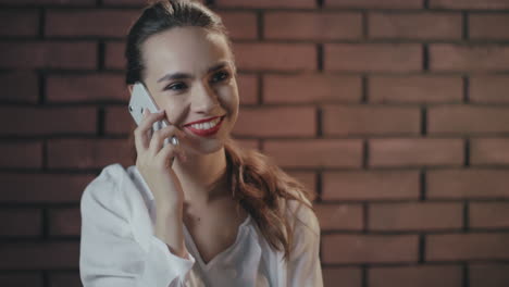 Bella-Mujer-Hablando-Por-Teléfono-Inteligente-Y-Sonriendo-En-El-Fondo-De-La-Pared-De-Ladrillo