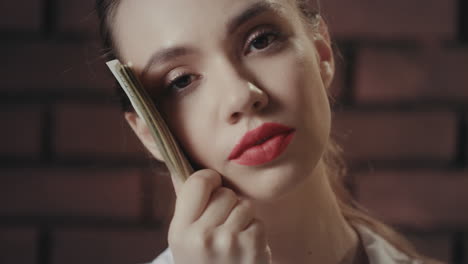 Thinking-woman-holding-money-stack-near-face-and-looking-into-camera