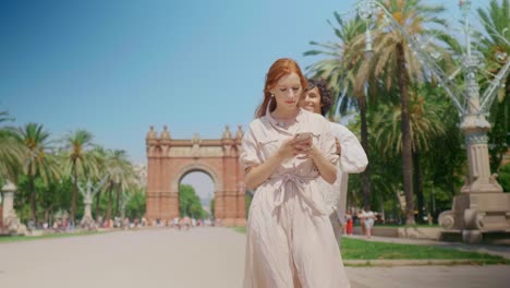 Focused-girl-looking-mobile-phone-near-monument.-Smiling-man-closing-woman-eyes