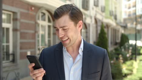 Closeup-businessman-smiling-with-phone-at-street.-Man-typing-message-outdoor