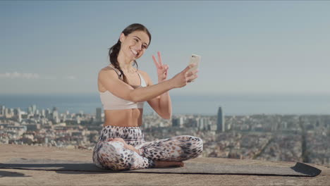 Sportsoman-taking-selfie-on-smartphone-at-city.-Yoga-woman-sitting-in-lotus-pose