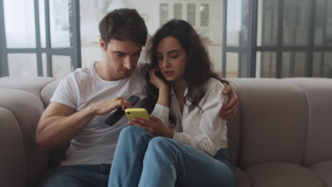 Happy-couple-sitting-on-sofa-with-mobile-phones-together.-Smiling-man-and-woman