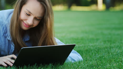 Happy-woman-laughing-while-using-laptop-computer-on-green-lawn-in-summer-park