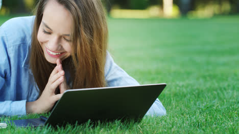 Smiling-woman-lying-on-green-grass-and-typing-on-laptop-in-summer-park