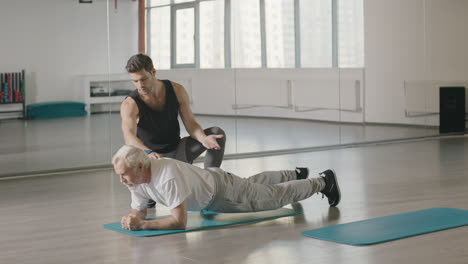 Aged-man-training-plank-workout-with-sport-coach-in-fitness-club-together