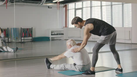Pensioner-man-doing-stretching-exercise-with-fitness-instructor-in-gym-together