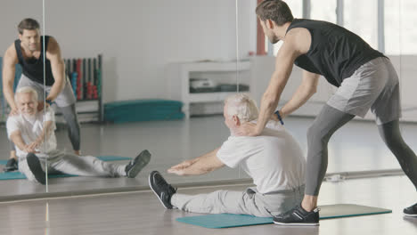 Sport-coach-helping-retired-man-doing-stretch-exercise-in-fitness-club.