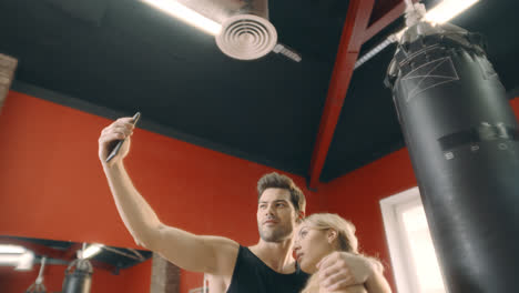 Cheerful-couple-hugging-for-mobile-selfie-in-fitness-gym.