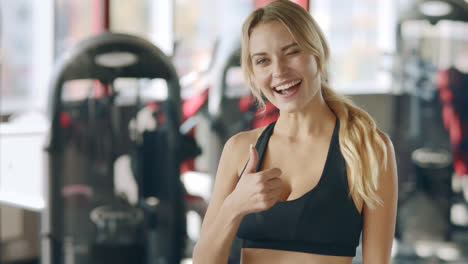 Fitness-woman-showing-thumb-up-and-winking-eyes-on-sport-club-background