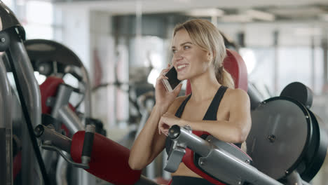 Cheerful-woman-talking-mobile-phone-at-fitness-training-machine-in-sport-gym.