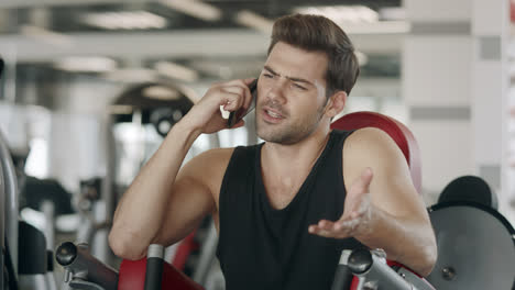 Athlete-man-talking-smartphone-after-training-on-sport-machine-in-fitness-gym