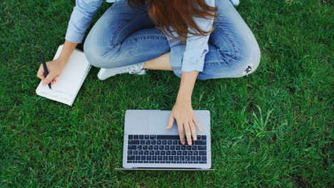 Woman-student-working-on-laptop-computer-and-writing-notes-in-notebook-in-park