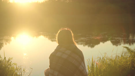 Lonely-girl-in-plaid-sitting-on-river-bank-in-rays-of-departing-sun.-Lonely-woman