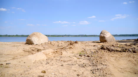 Sand-and-huge-stones-on-shore-of-lake-near-village.-Butterflies-fly-near-lake
