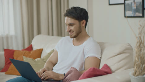 Smiling-man-working-with-laptop-computer-at-home-sofa.-Young-man-chatting-online