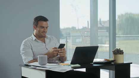 Business-man-scrolling-mobile-phone-at-work-place.-Smiling-man-having-break.