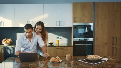 Concentrated-man-working-at-luxury-kitchen.-Smiling-wife-talking-with-husband