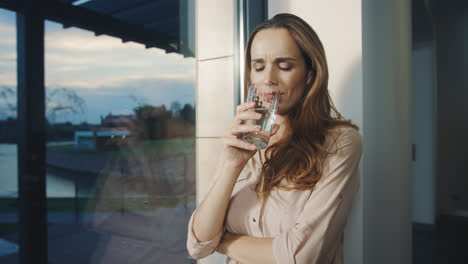 Mujer-Relajada-Bebiendo-Agua-Cerca-De-La-Ventana-Panorámica.-Señora-Sedienta-Bebiendo-Agua
