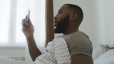 Shocked-black-man-sitting-in-bed.-Smartphone-falling-out-of-man-hand.