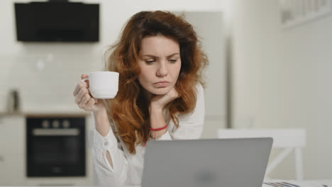 Focused-woman-working-laptop-at-open-kitchen.-Young-lady-drinking-coffee