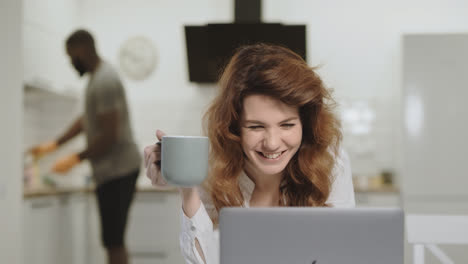 Smiling-white-woman-reading-happy-news-at-laptop-in-morning-kitchen.