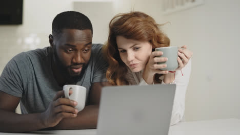 Tired-black-man-looking-laptop-screen-at-home.-Woman-bringing-tea-to-guy.