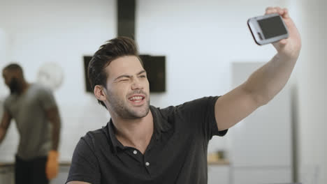 Smiling-man-making-selfie-on-phone-at-open-kitchen.-Young-guy-holding-phone.