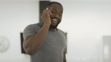 Hombre-Negro-Sonriente-Hablando-Por-Teléfono-En-Casa.-Chico-Joven-De-Pie-Con-Teléfono-Inteligente