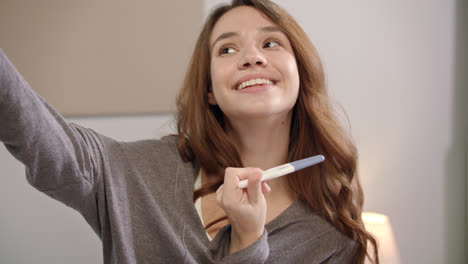 Happy-woman-taking-selfie-photo-with-pregnancy-test-on-mobile-phone-at-bedroom